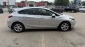 Silver four-door hatchback car parked in a paved lot with other vehicles and storefronts in the background (side view).