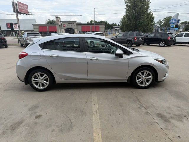Silver four-door hatchback car parked in a paved lot with other vehicles and storefronts in the background (side view).