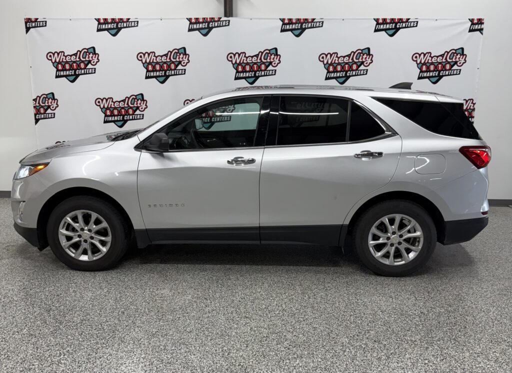 Side view of a silver Chevrolet Traverse SUV parked indoors with a Wheel City Auto Finance Centers banner in the background.