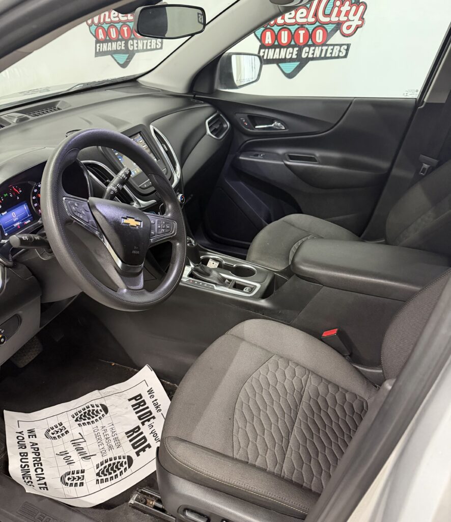 Driver's seat interior of a Chevrolet SUV, steering wheel with logo, dashboard, center console, and a paper floor mat on the driver side