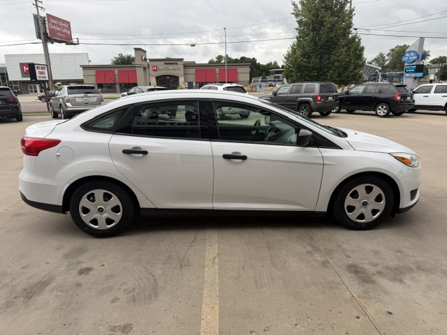 Side view of a white four‑door sedan parked in a car dealership lot, with other vehicles and storefronts in the background.