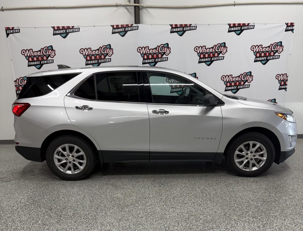 Side view of a silver Chevrolet Equinox SUV indoors with a repeating Wheel City Auto Finance Centers banner in the background.