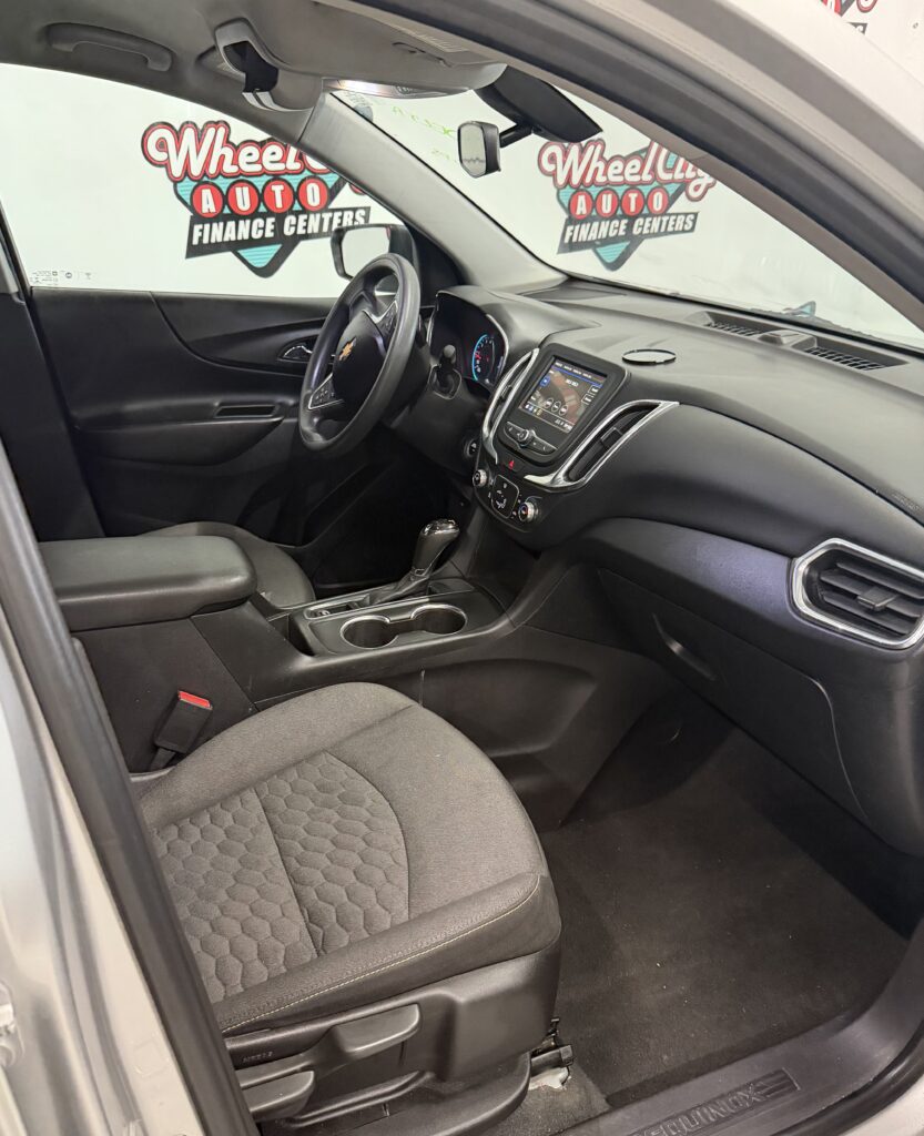 Front interior of a Chevrolet sedan showing the driver seat, steering wheel, center console with touchscreen, and gear shifter in a dark cloth interior.