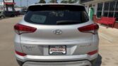 Rear view of a white Hyundai Tucson with 'Tucson' and 'Sport' badges, parked in front of a dealership; window sticker on the rear window.