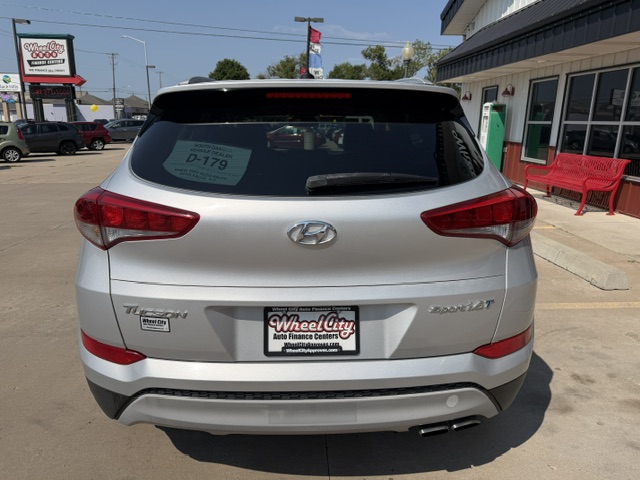 Rear view of a white Hyundai Tucson with 'Tucson' and 'Sport' badges, parked in front of a dealership; window sticker on the rear window.