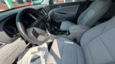 Front interior of a light gray SUV, showing the driver's seat, steering wheel, and center console from a passenger-side angle at a dealership. The photo captures the clean, light upholstery, gear shifter, and dashboard controls against the car’s open door and large side window.