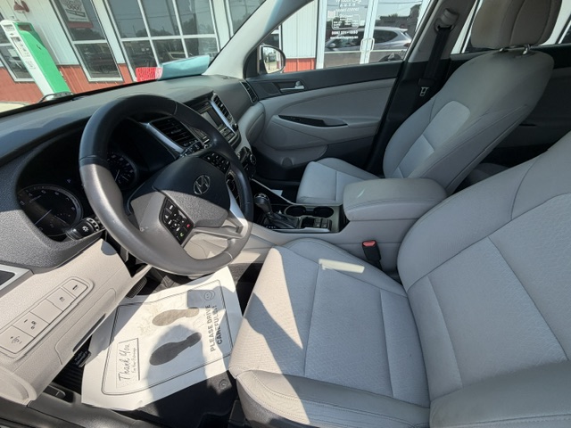 Front interior of a light gray SUV, showing the driver's seat, steering wheel, and center console from a passenger-side angle at a dealership. The photo captures the clean, light upholstery, gear shifter, and dashboard controls against the car’s open door and large side window.