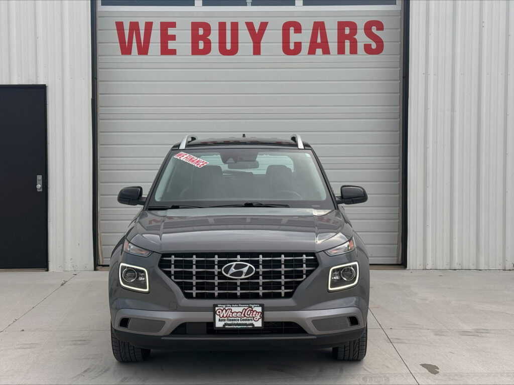 Front view of a gray Hyundai SUV parked in front of a metal building with a red 'WE BUY CARS' sign on the garage door, windshield sticker visible.