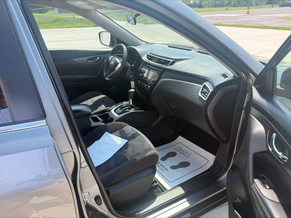 Interior of a parked car viewed from the open door: steering wheel, center console, and dashboard visible with papers on the front passenger seat.
