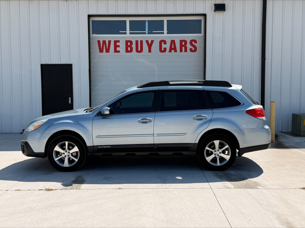 Silver SUV with roof rails parked in front of a white metal building featuring a large garage door that reads 'WE BUY CARS' in red letters.