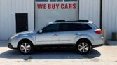 Silver SUV with roof rails parked in front of a white metal building featuring a large garage door that reads 'WE BUY CARS' in red letters.