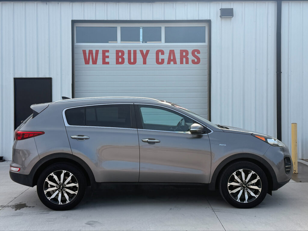 Car dealership with a large 'WE BUY CARS' sign on a garage door; silver SUV in foreground.