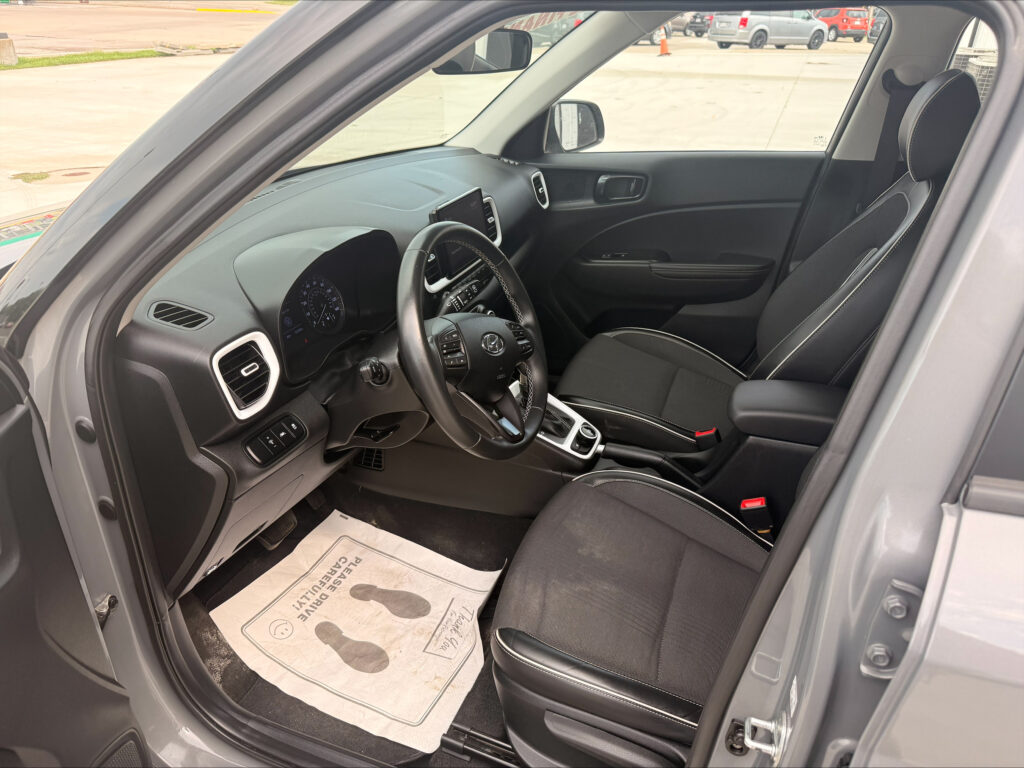 Interior of a gray SUV front cabin with steering wheel, dashboard, center console, and black-stitched seats; paper floor mat visible on driver side