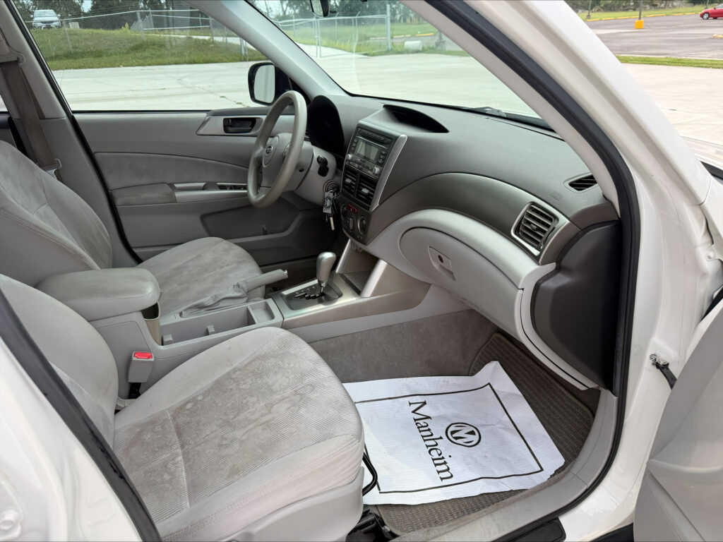 Interior of a light gray car: front seats, steering wheel, center console, and dashboard visible from the open driver-side door.",