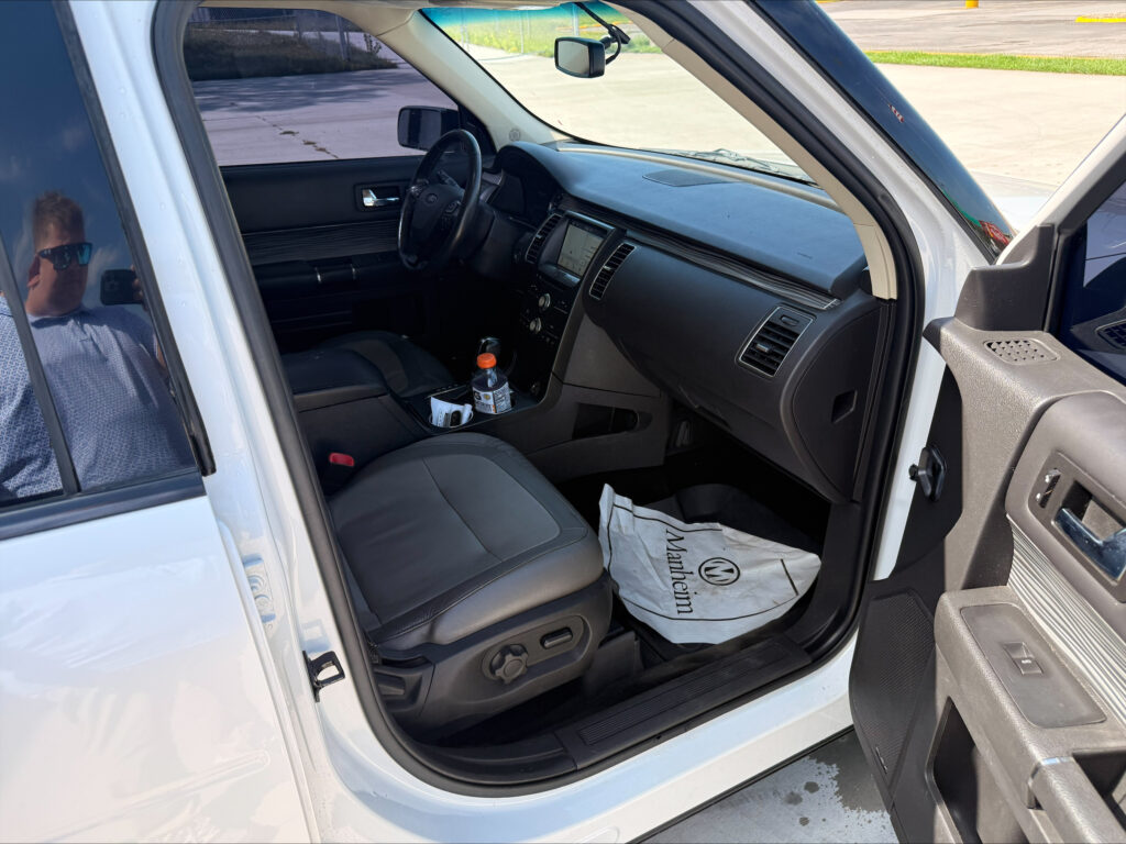 Inside a white SUV showing the driver's seat and dashboard; a bottle and papers sit on the floor, with a man wearing sunglasses reflected in the window.