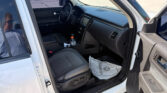 Inside a white SUV showing the driver's seat and dashboard; a bottle and papers sit on the floor, with a man wearing sunglasses reflected in the window.