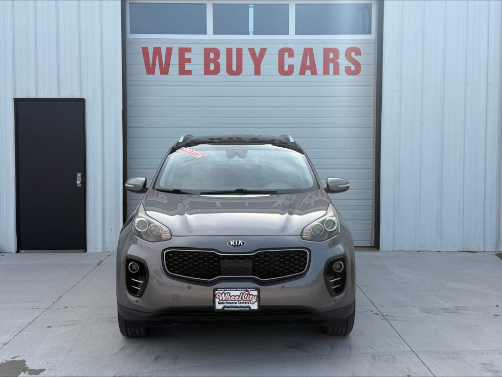 Gray Kia SUV parked in front of a garage door with a large red 'WE BUY CARS' sign above it.