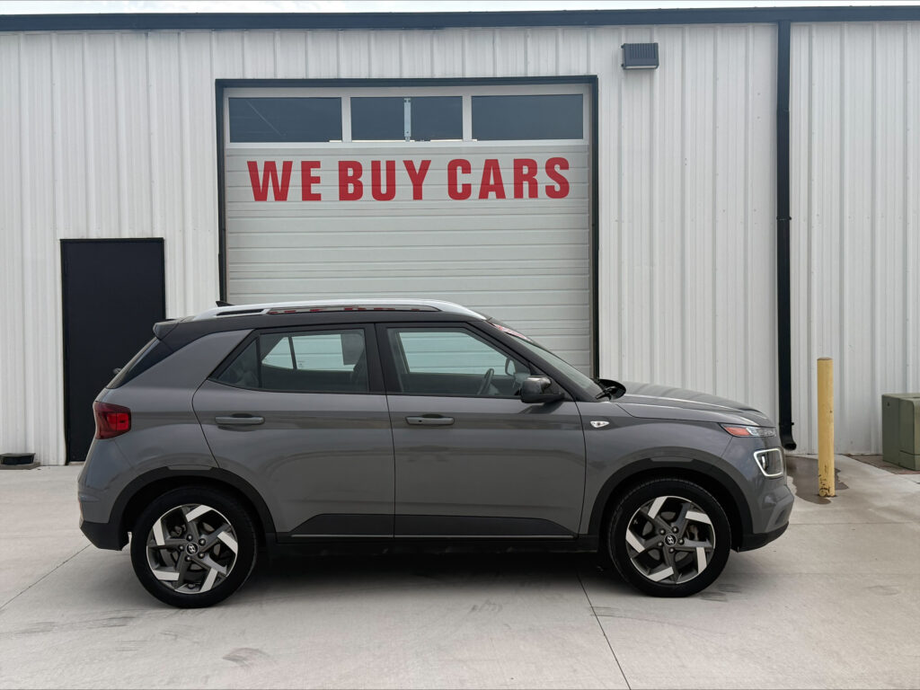 Gray SUV parked in front of a white metal building with a large garage door that says 'WE BUY CARS' in red letters.