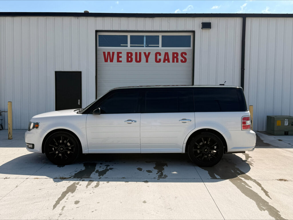 White minivan parked in front of a building with a large 'WE BUY CARS' sign on a garage door in the background.