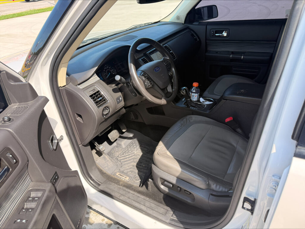 Interior view from the driver’s side showing the steering wheel, dashboard, center console, and a bottle on the floor mat near the pedals (Ford).