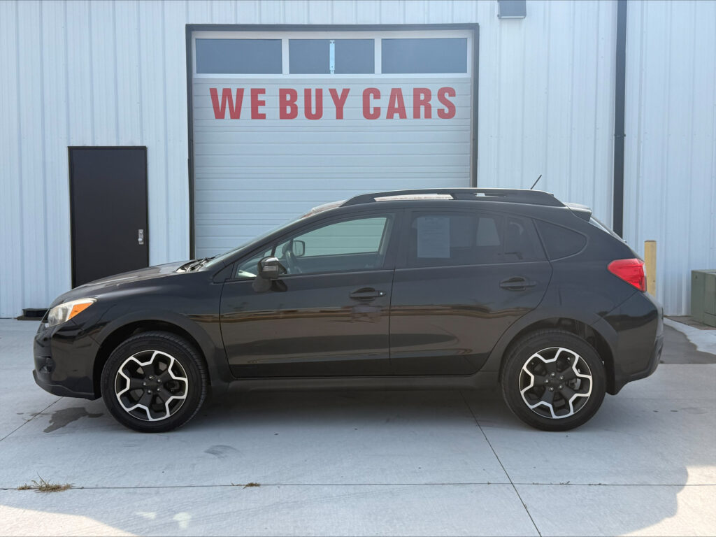 Black SUV parked in front of a light blue industrial building with a large garage door that reads 'WE BUY CARS' in red letters, daytime.