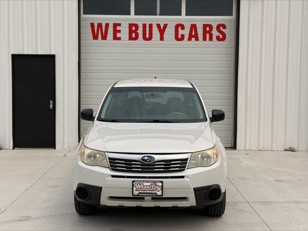 Front view of a white SUV parked in front of a metal building with a large red sign saying WE BUY CARS above the garage door.
