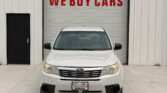 Front view of a white SUV parked in front of a metal building with a large red sign saying WE BUY CARS above the garage door.