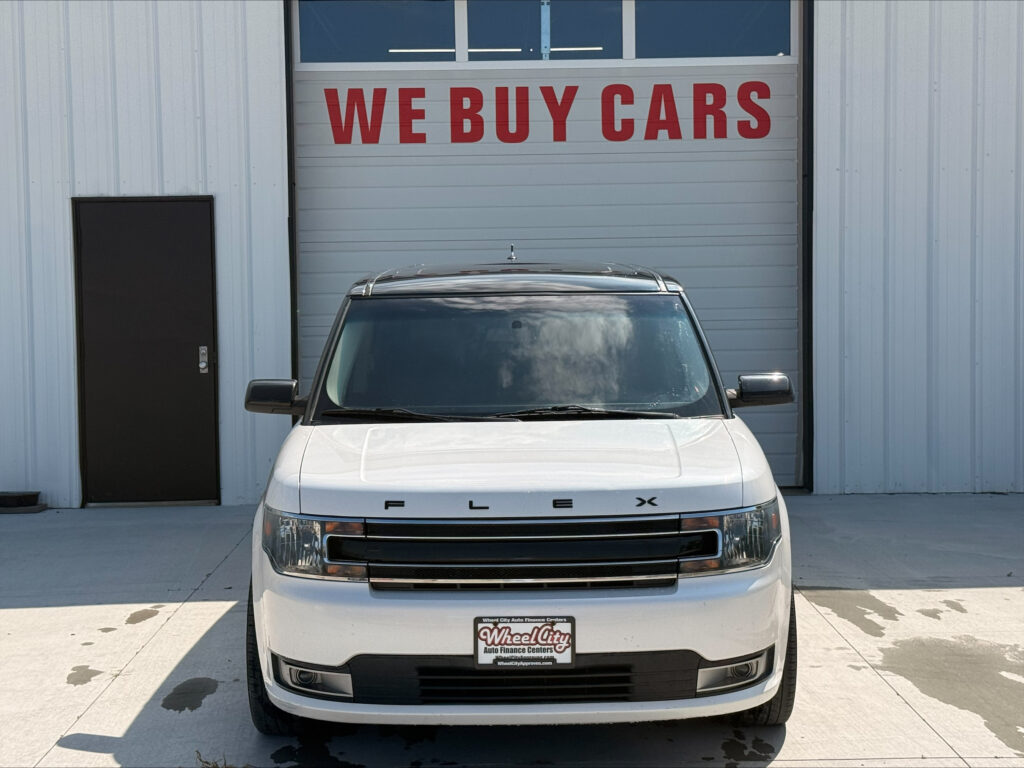 Front view of a white Ford Flex parked in front of a building with a large red 'WE BUY CARS' sign on the garage door.