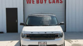 Front view of a white Ford Flex parked in front of a building with a large red 'WE BUY CARS' sign on the garage door.