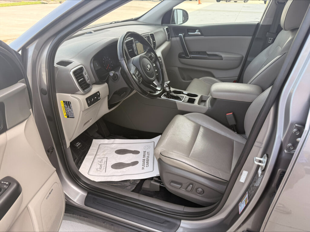 Interior of a car viewed from the open driver’s door: steering wheel, dashboard, center console, and front passenger seat visible in gray upholstery. The floor has a paper mat.