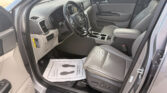 Interior of a car viewed from the open driver’s door: steering wheel, dashboard, center console, and front passenger seat visible in gray upholstery. The floor has a paper mat.