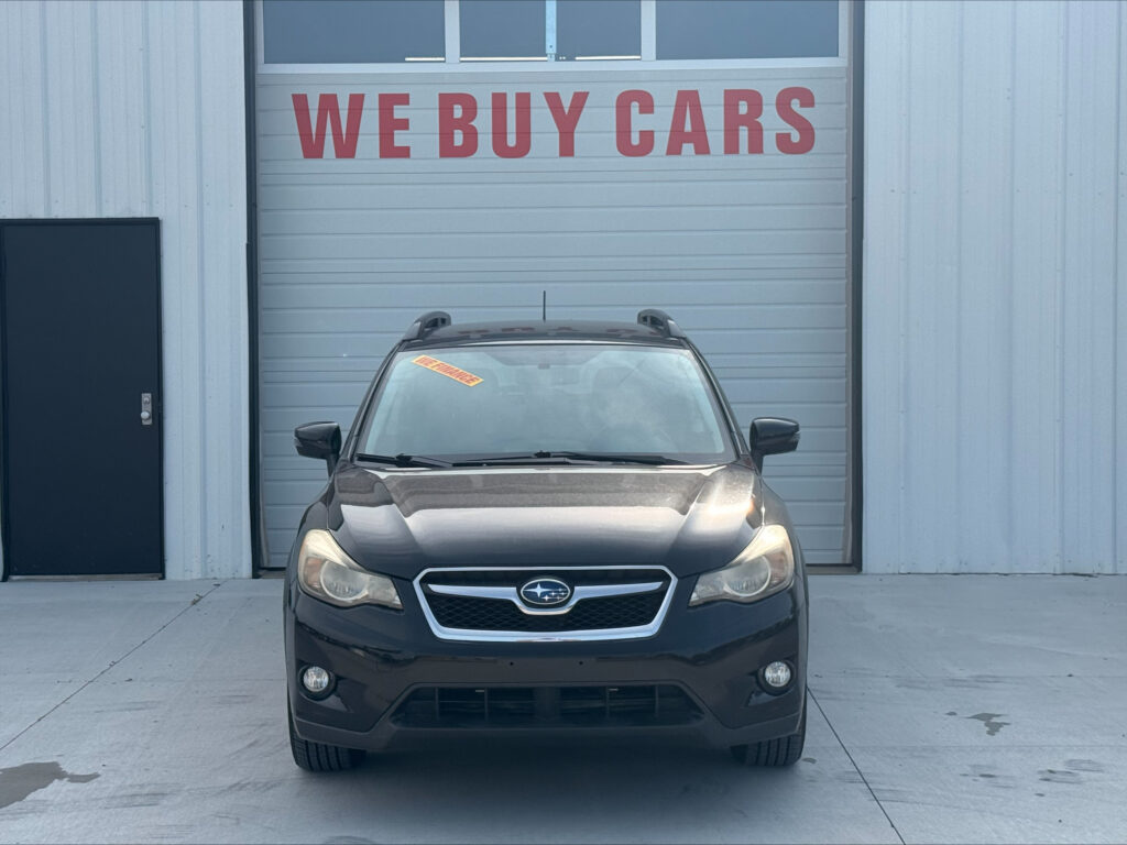 Front view of a dark gray Subaru SUV parked in front of a large light-gray garage door with a red 'WE BUY CARS' sign above the door.