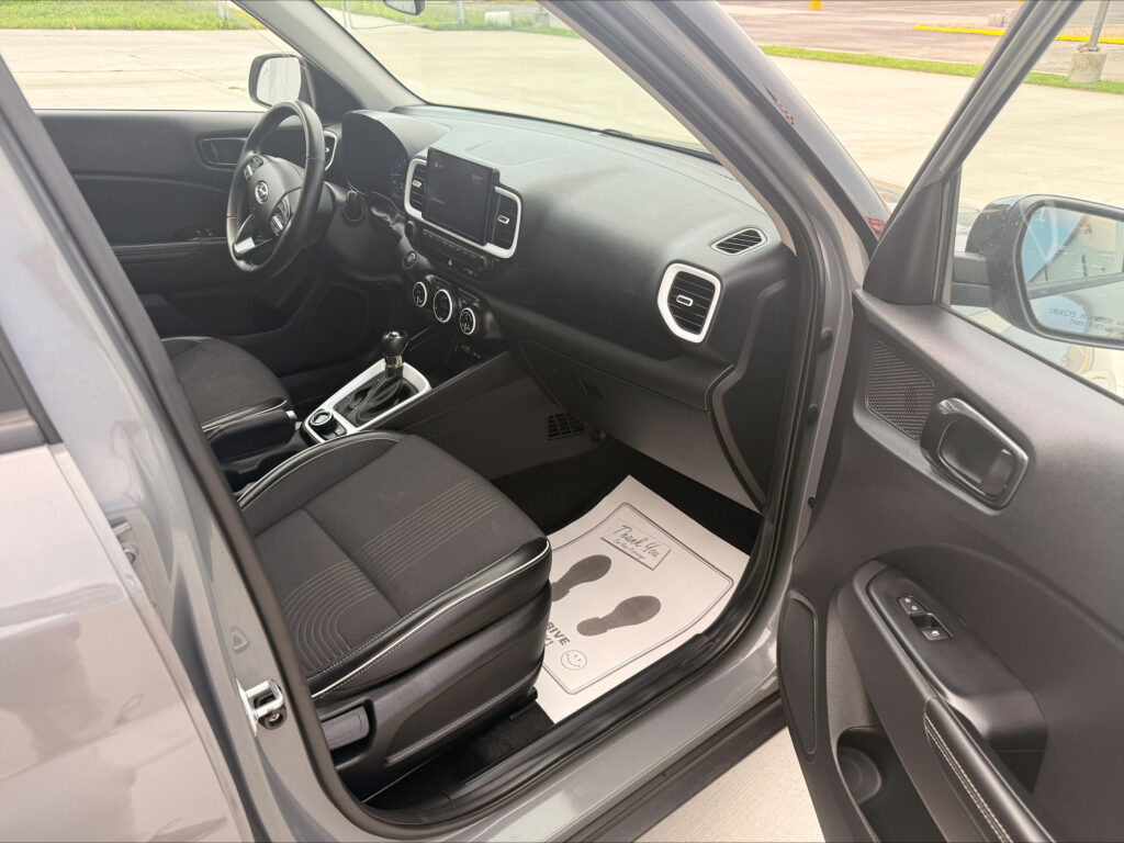 Interior of a car with the driver's seat, steering wheel, center touchscreen, gear shifter, and a floor mat that says 'Thank You'.