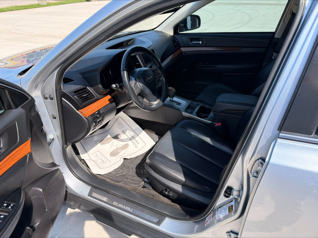 Open driver's door view of a Subaru interior, showing the steering wheel, dashboard, center console, and a paper floor mat on the driver’s side