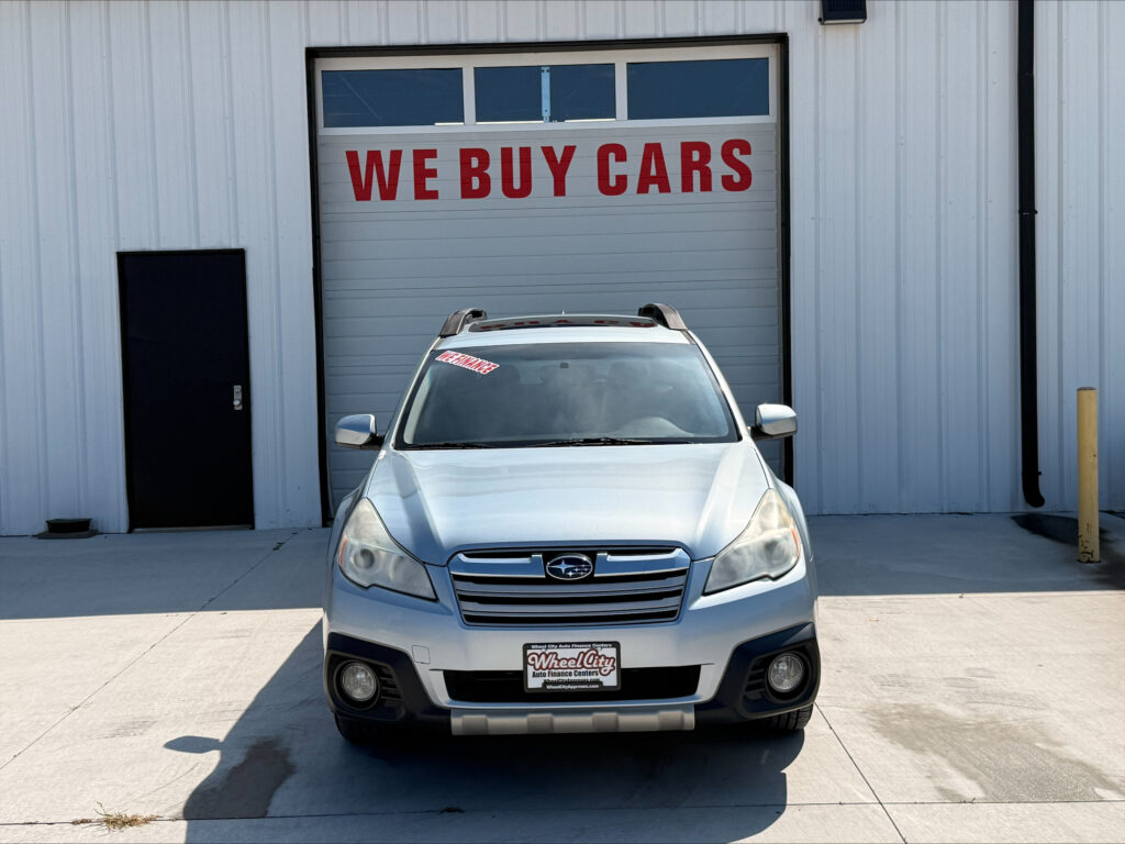 Silver SUV parked in front of a garage door with a large red 'WE BUY CARS' sign above it; car has a Wheel City license plate frame and dealership sticker visible on windshield
